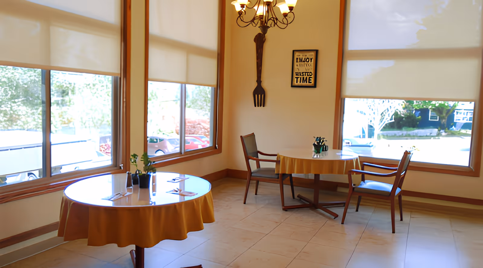 A bright dining area with large windows covered by white roller shades. Two round tables with yellow tablecloths are set with utensils and small potted plants. Wooden chairs with cushions are placed around the tables. A decorative wall piece shaped like a large fork and a framed sign hang on the wall. A chandelier with multiple lights hangs from the ceiling.
