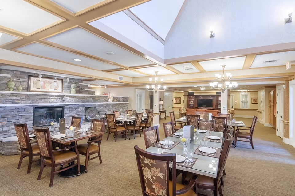 Spacious, well-lit dining room with multiple set tables and upholstered chairs, a stone fireplace wall, and skylights.