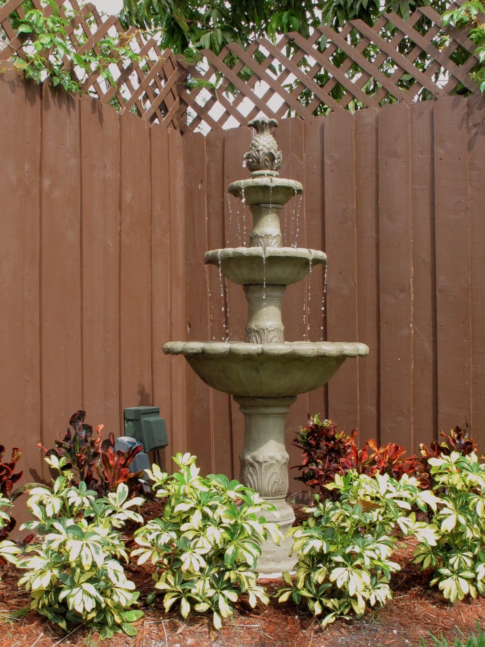 A three-tiered stone water fountain with water flowing from the top tier to the bottom, surrounded by green and red leafy plants, set against a brown wooden fence with lattice work at the top and some green vines growing over it.