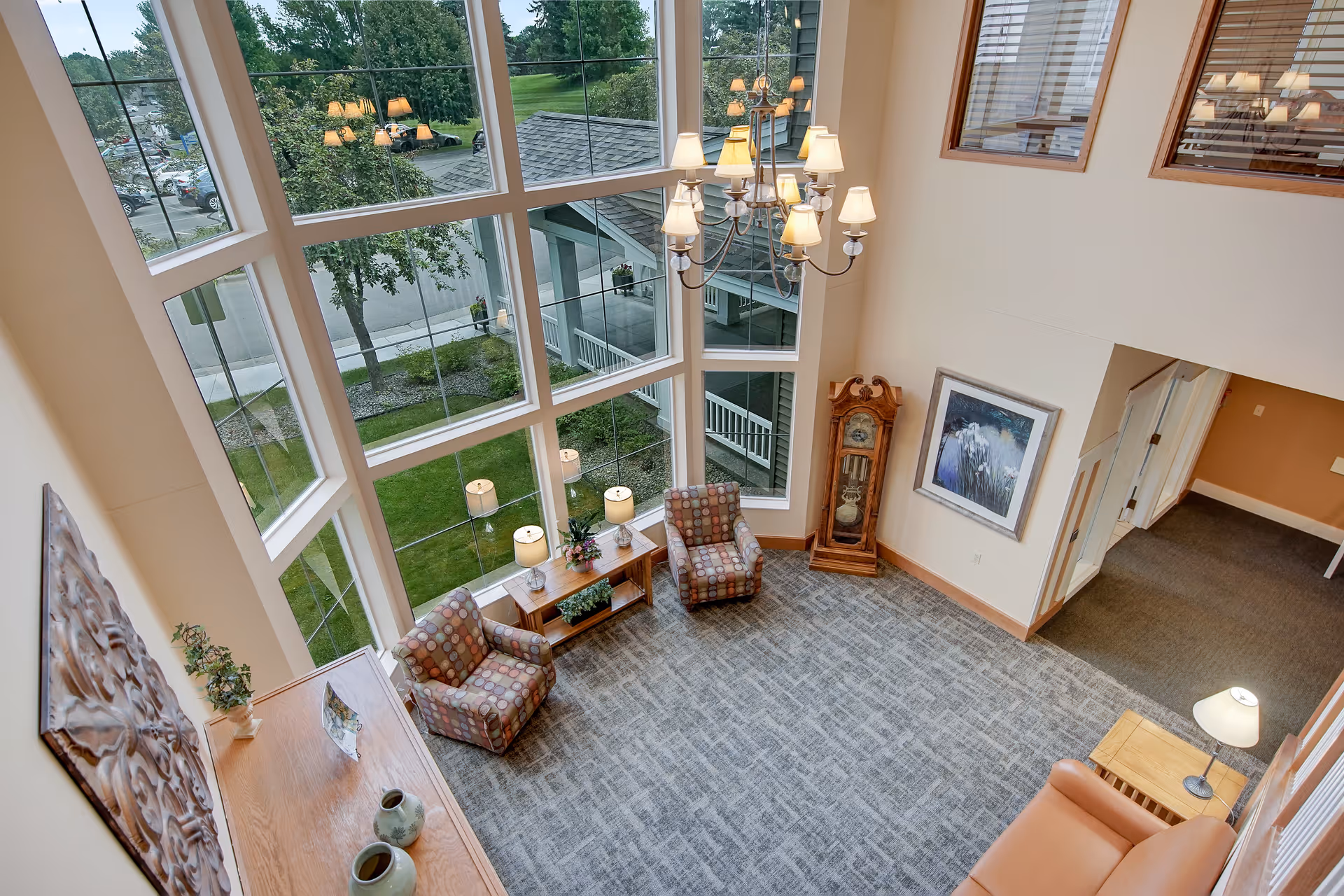 View from above of a spacious living room area with large floor-to-ceiling windows showing an outdoor green space. The room features two patterned armchairs, a wooden console table with lamps and plants, a grandfather clock, a framed painting on the wall, a chandelier hanging from the ceiling, and a small side table with a lamp next to a leather sofa.