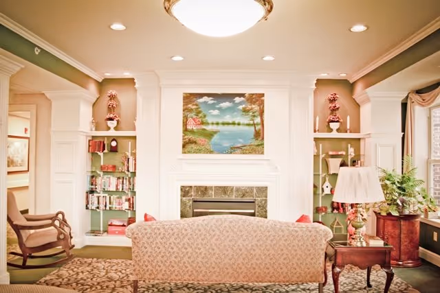 A cozy living room area with a floral patterned sofa facing a fireplace. Above the fireplace is a painting of a serene lake scene with trees and a small red house. On either side of the fireplace are built-in shelves with books, decorative items, and candles. To the left is a wooden rocking chair, and to the right is a side table with a lamp and a potted plant. The room has soft lighting from ceiling fixtures and a large window with curtains on the right side.