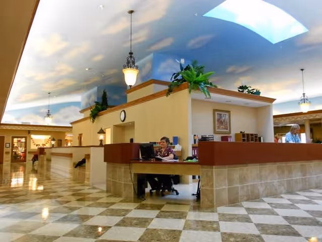Interior view of a senior living facility reception area with a checkered tile floor, beige walls, and a ceiling painted with a blue sky and clouds. Two staff members are seated at desks behind a counter, with plants and framed artwork decorating the space.