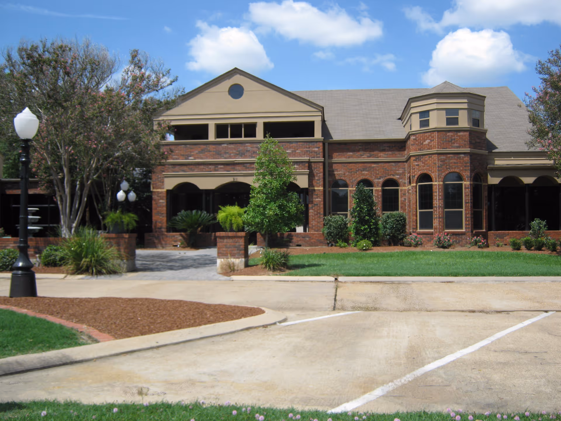 Front exterior of a brick two-story building with arched windows, landscaped shrubs, and a parking area under a blue sky.