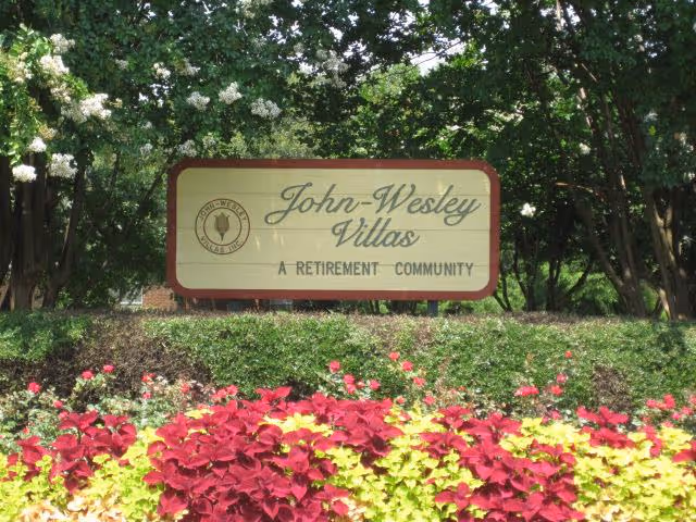 A large sign for John-Wesley Villas, a retirement community, surrounded by green bushes and colorful red and yellow flowers, with trees in the background.