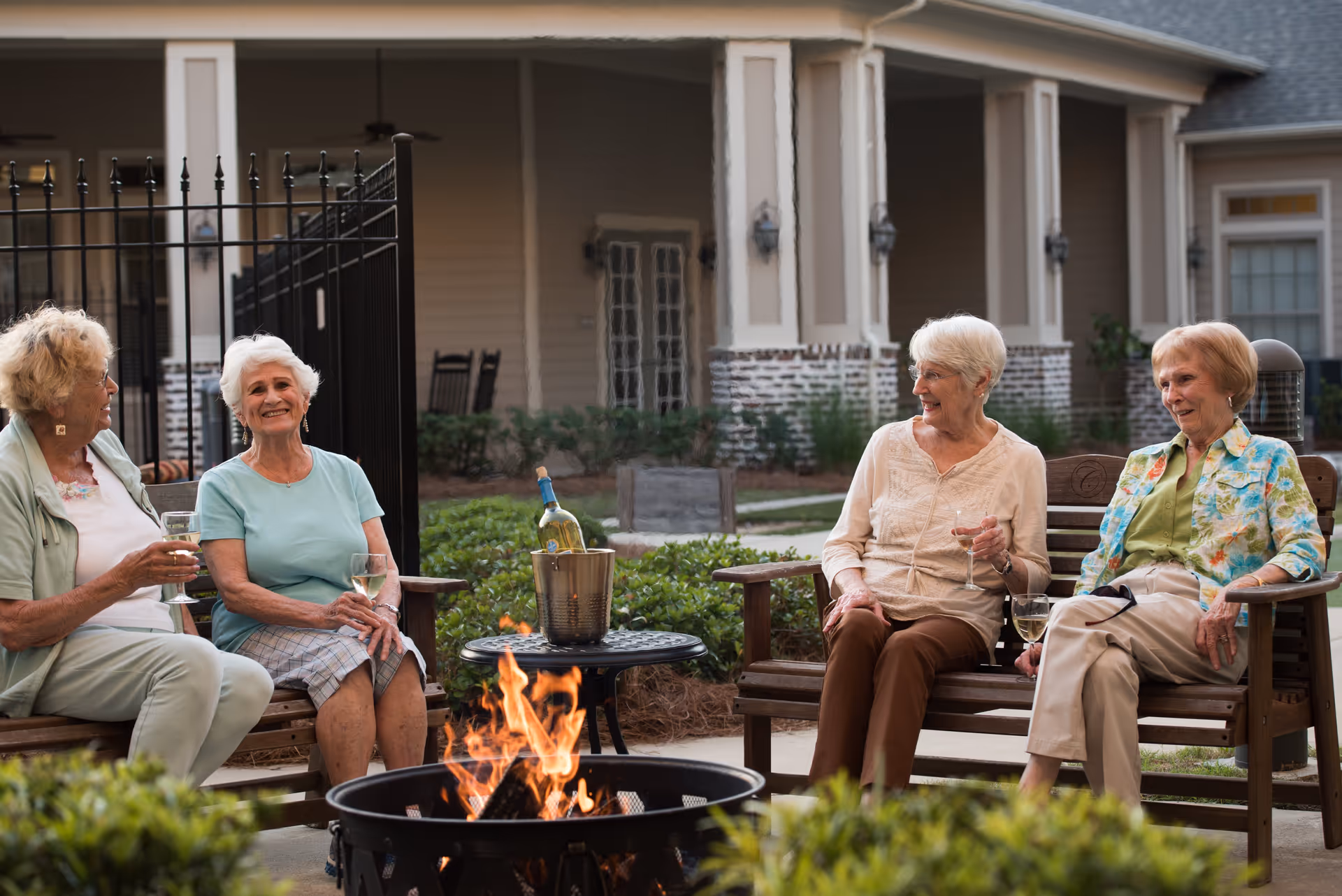 Four elderly women sitting on wooden benches around a fire pit outdoors, enjoying drinks and smiling, with a building and greenery in the background.