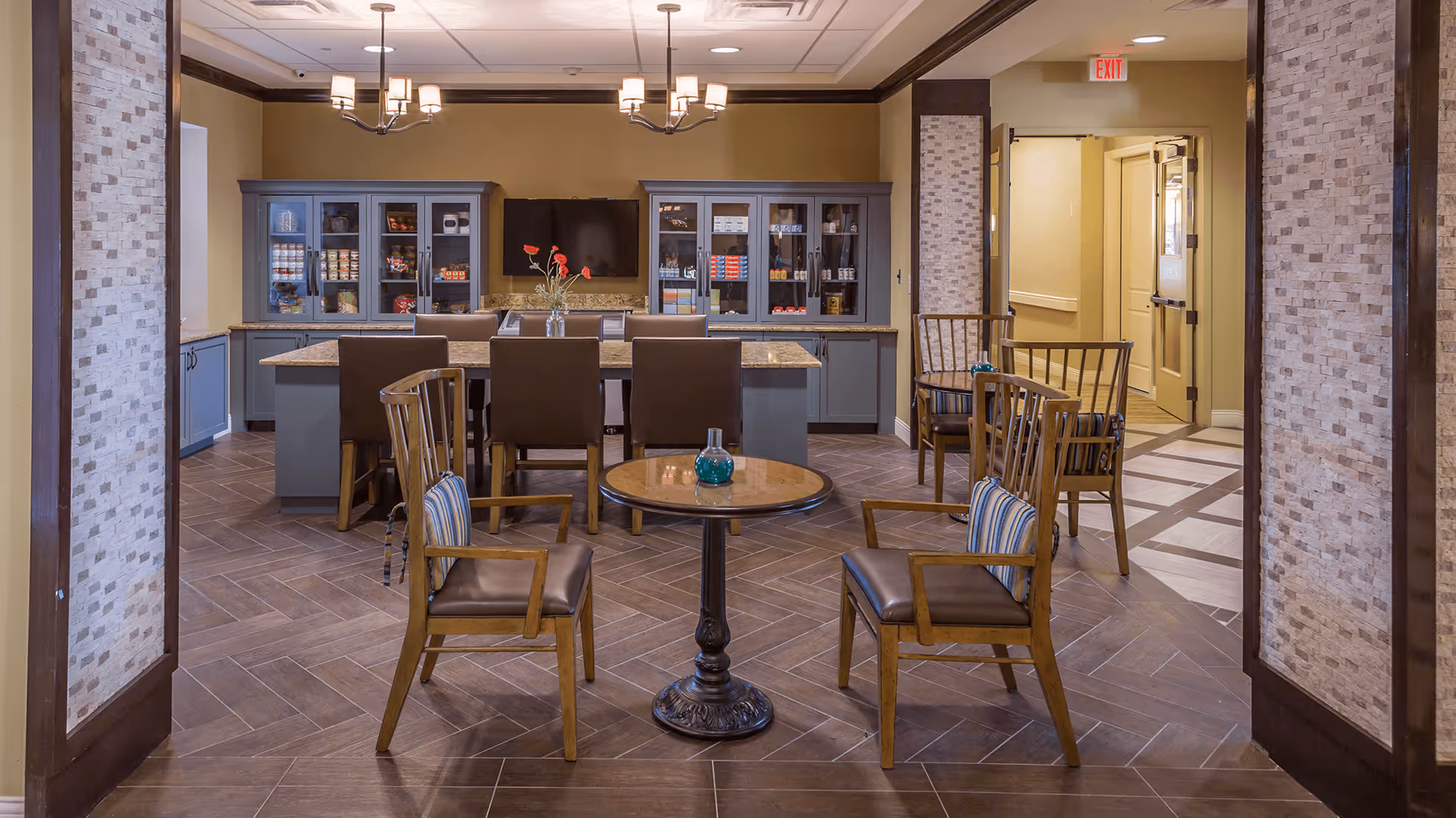 A cozy common area with a small round table and two wooden chairs with cushions in the foreground. In the background, there is a larger rectangular table with six chairs and a cabinet with glass doors displaying various items. The room has warm lighting with two chandeliers hanging from the ceiling and a hallway leading to a door with an exit sign above it.