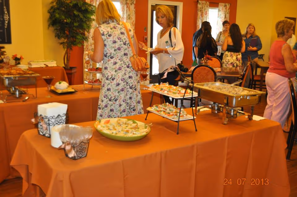 A buffet table covered with an orange tablecloth displaying various appetizers and dishes in a dining area. Several women are standing around, some serving themselves food and others conversing. The room has warm yellow walls, floral curtains, and wooden chairs with orange cushions.