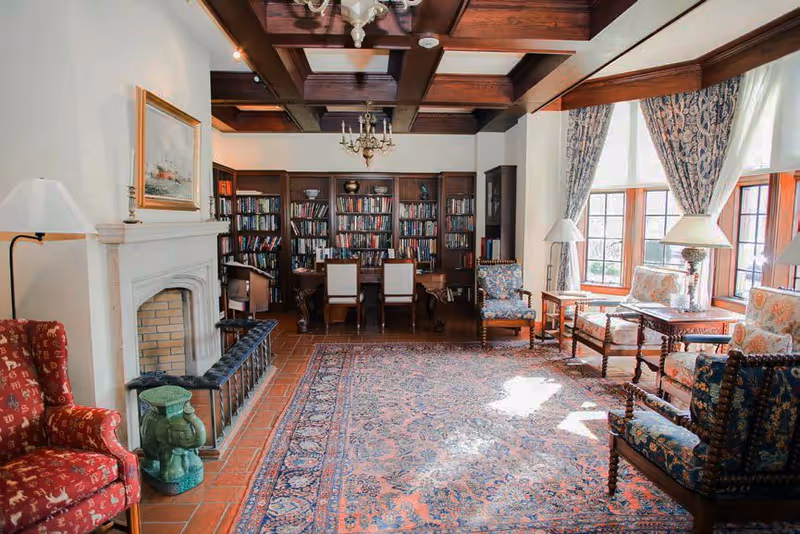 Sunlit elegant sitting room with a fireplace, large patterned rug, multiple upholstered chairs and side tables, and a bookshelf-lined wall.