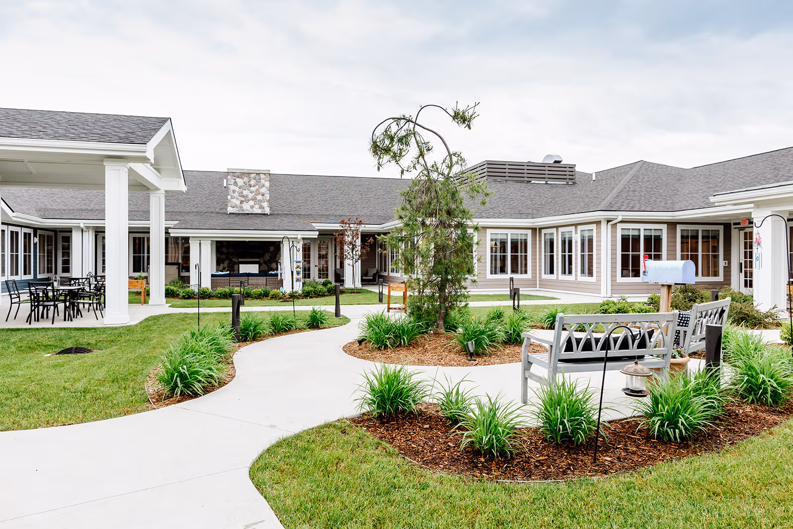 Landscaped courtyard with curved walking paths, benches, and a covered patio in front of a single-story memory care building.