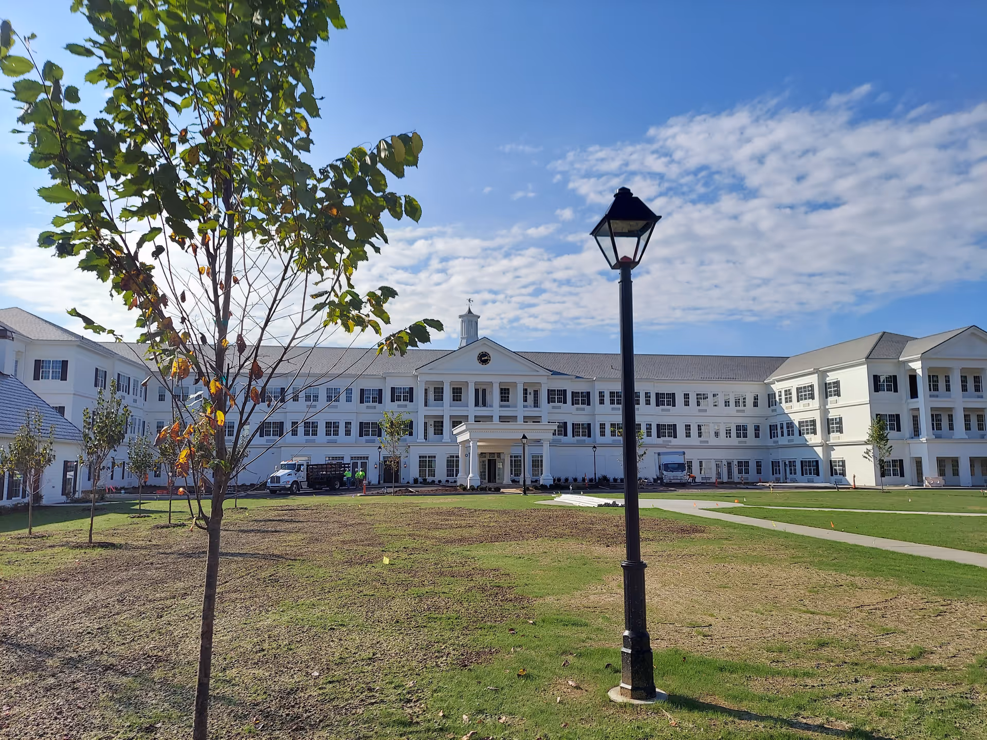 Exterior view of a large, white, three-story senior living facility building with many windows and a central entrance with columns. There is a grassy area with young trees and a black lamppost in the foreground under a partly cloudy blue sky.