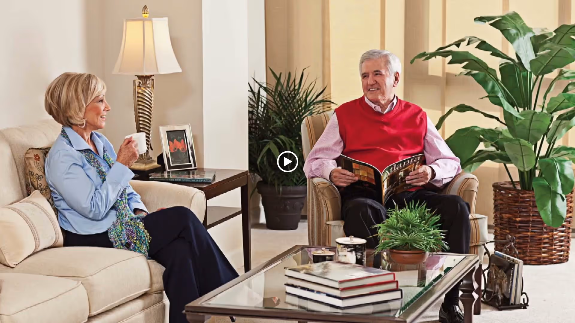 An elderly woman and man sitting in a well-lit living room. The woman is seated on a beige sofa holding a white cup, while the man sits in an armchair holding a magazine. The room features a glass coffee table with stacked books and a potted plant, a side table with a lamp and framed picture, and large green plants in the background.