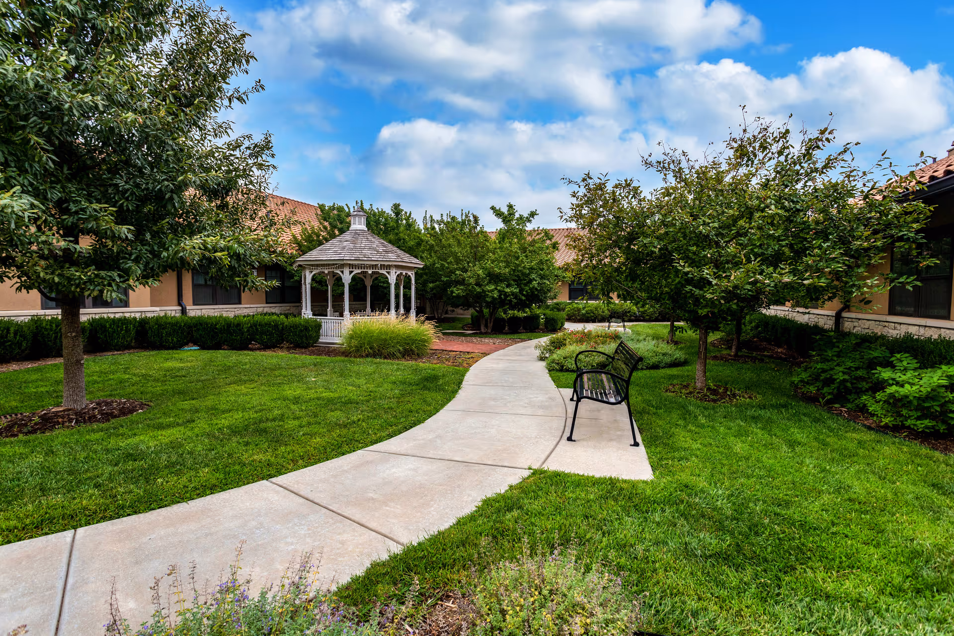 A well-maintained outdoor garden area with a curved concrete pathway, green grass, trees, shrubs, a black metal bench, and a white gazebo under a partly cloudy blue sky.