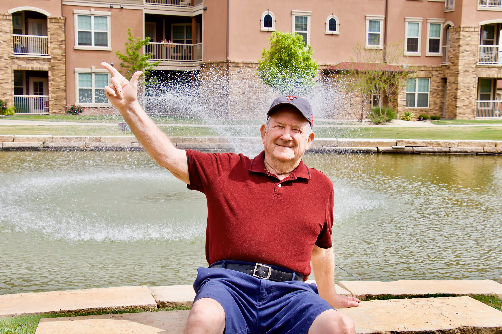 An elderly man in a red polo and blue shorts sits by a pond with a fountain and apartment buildings behind him, smiling and waving.