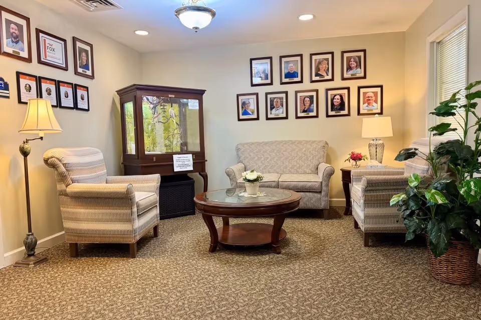 A cozy sitting area with two striped armchairs, a patterned loveseat, a round wooden coffee table with a glass top, and two table lamps. The walls are adorned with framed photographs and certificates. There is a large potted plant near a window with blinds, and a wooden display cabinet in the corner.
