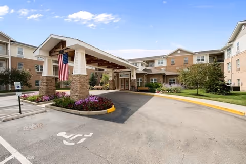 Front entrance of a senior living building with a covered porte-cochère, American flag, landscaping, and a driveway.