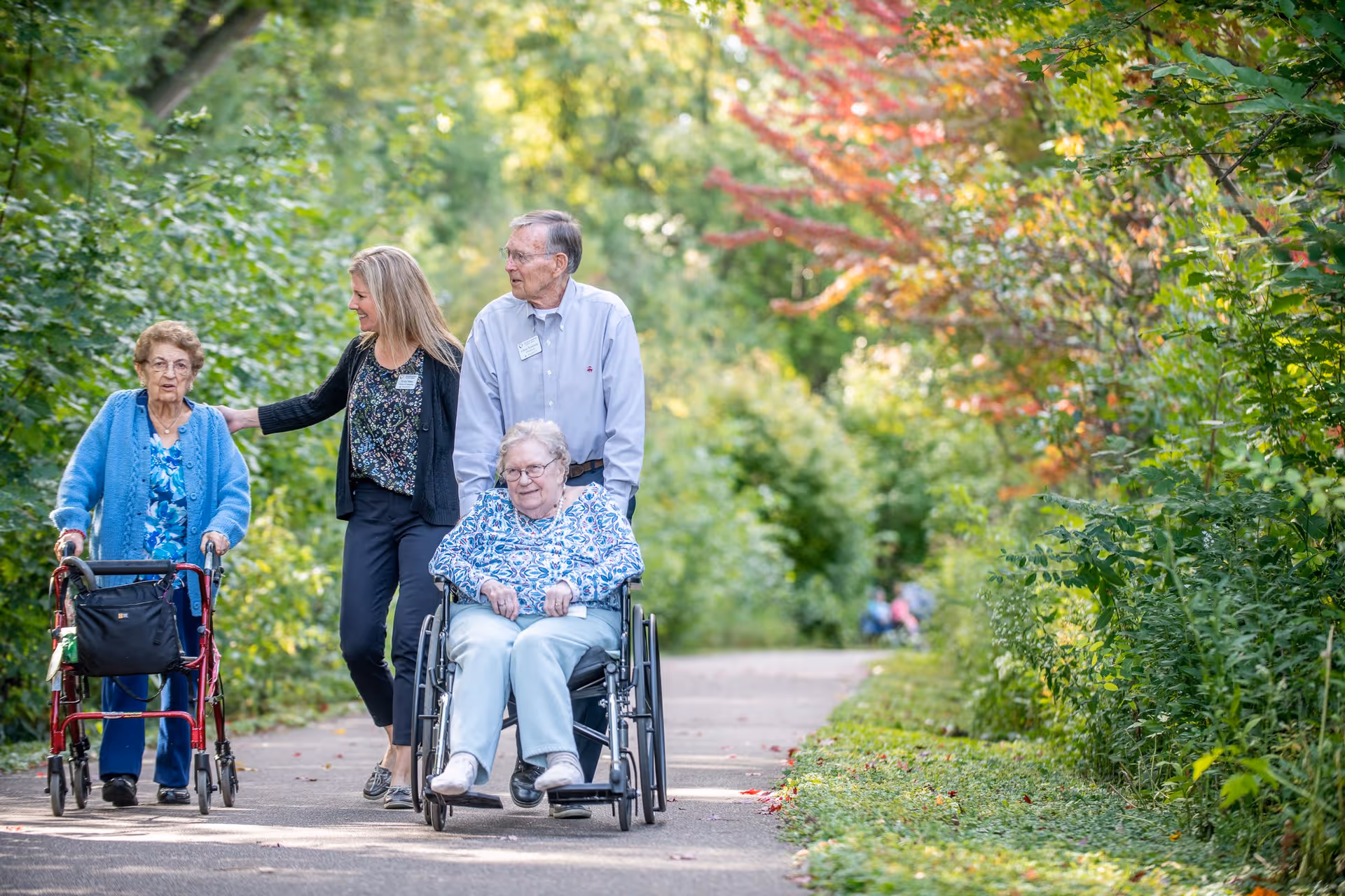 Two elderly women, one using a walker and the other in a wheelchair, are accompanied by a man and a woman walking with them on a paved path surrounded by lush green trees and foliage.