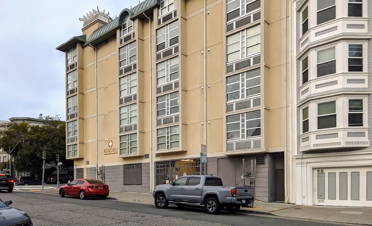 Exterior view of a multi-story beige building with large windows and a sign that reads Kokoro Assisted Living Inc. Several cars are parked along the street in front of the building.