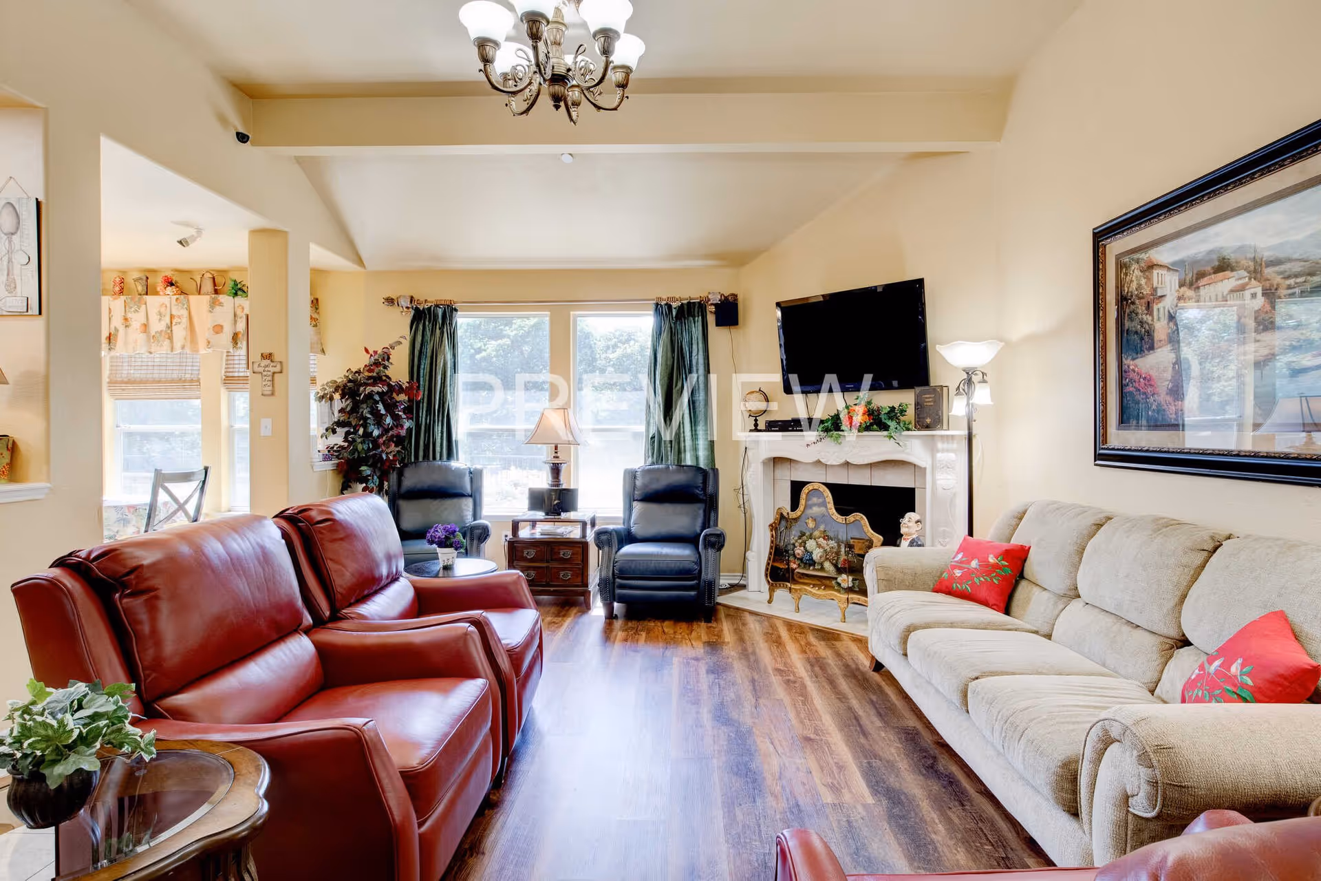 A cozy living room with wooden flooring, featuring two red leather recliners on the left, a beige sofa with red floral pillows on the right, and two black armchairs near large windows with green curtains. A white fireplace with a decorative screen and a mounted flat-screen TV is on the far wall. The room is decorated with a chandelier, a floor lamp, a side table with a plant, and a framed painting on the wall.