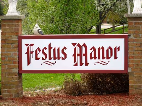 A Festus Manor sign between brick pillars with greenery in the background.