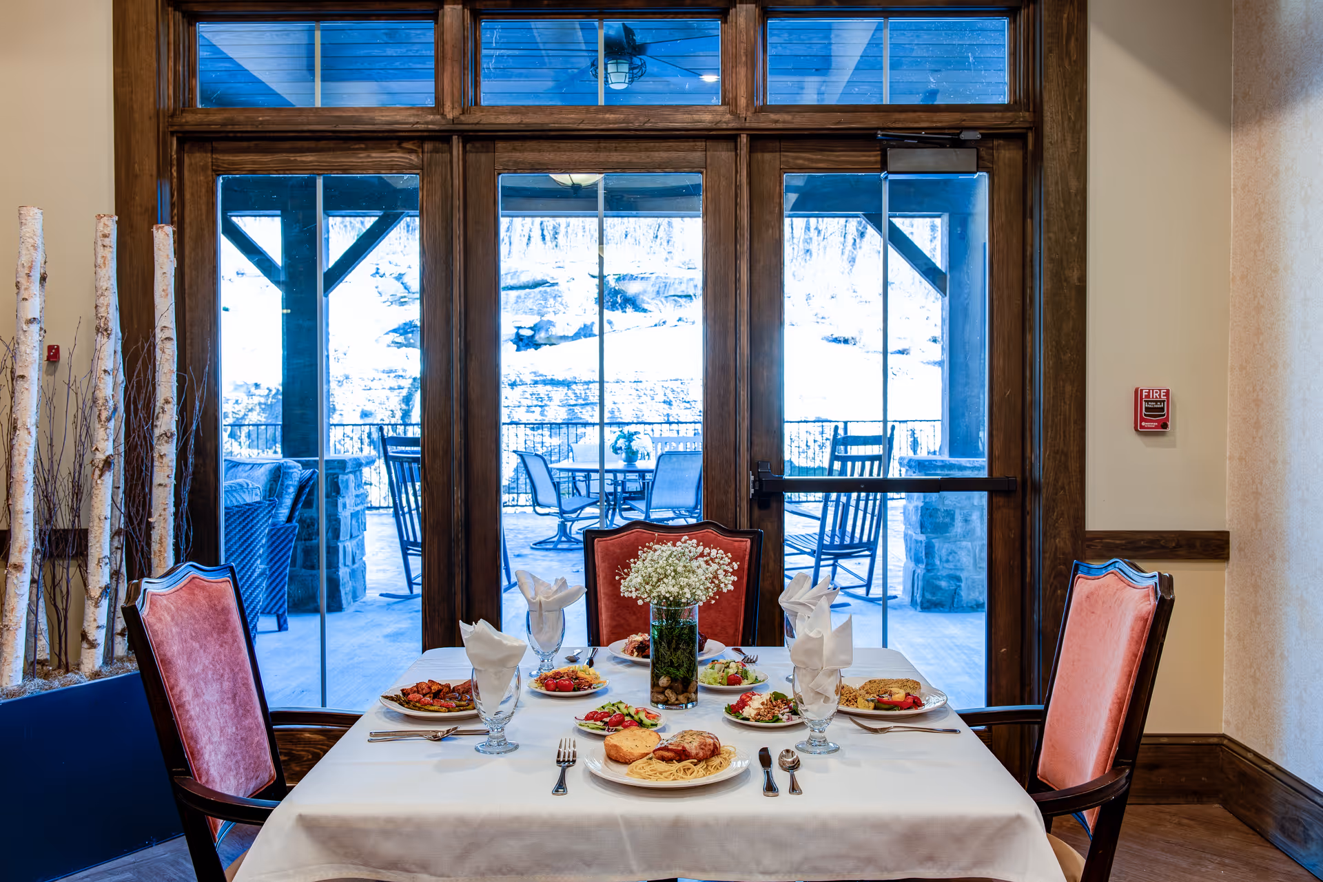 A dining table set for four with plates of food, glasses with folded napkins, and a vase with white flowers in the center. The table is positioned in front of large glass doors that open to an outdoor patio with chairs and tables. The room has wooden trim and a decorative arrangement of birch branches in a planter on the left side.