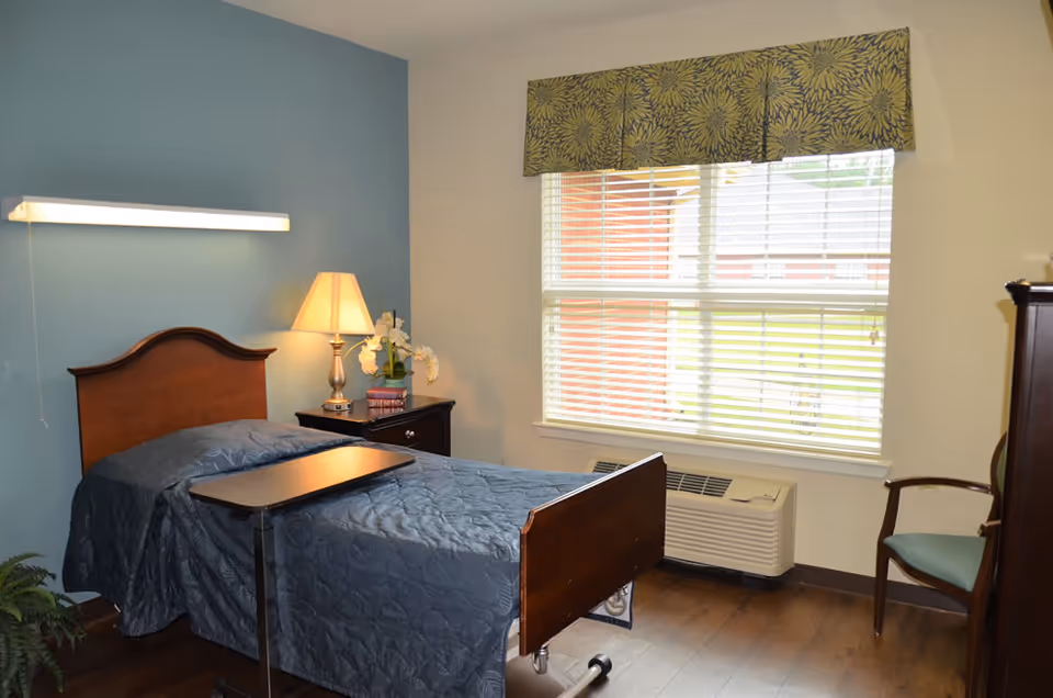 A senior care facility bedroom with a single bed covered in a dark blue quilt, a wooden headboard and footboard, a bedside table with a lamp and decorative flowers, a window with blinds and a patterned valance, a wooden chair with a cushion, and a small overbed table.