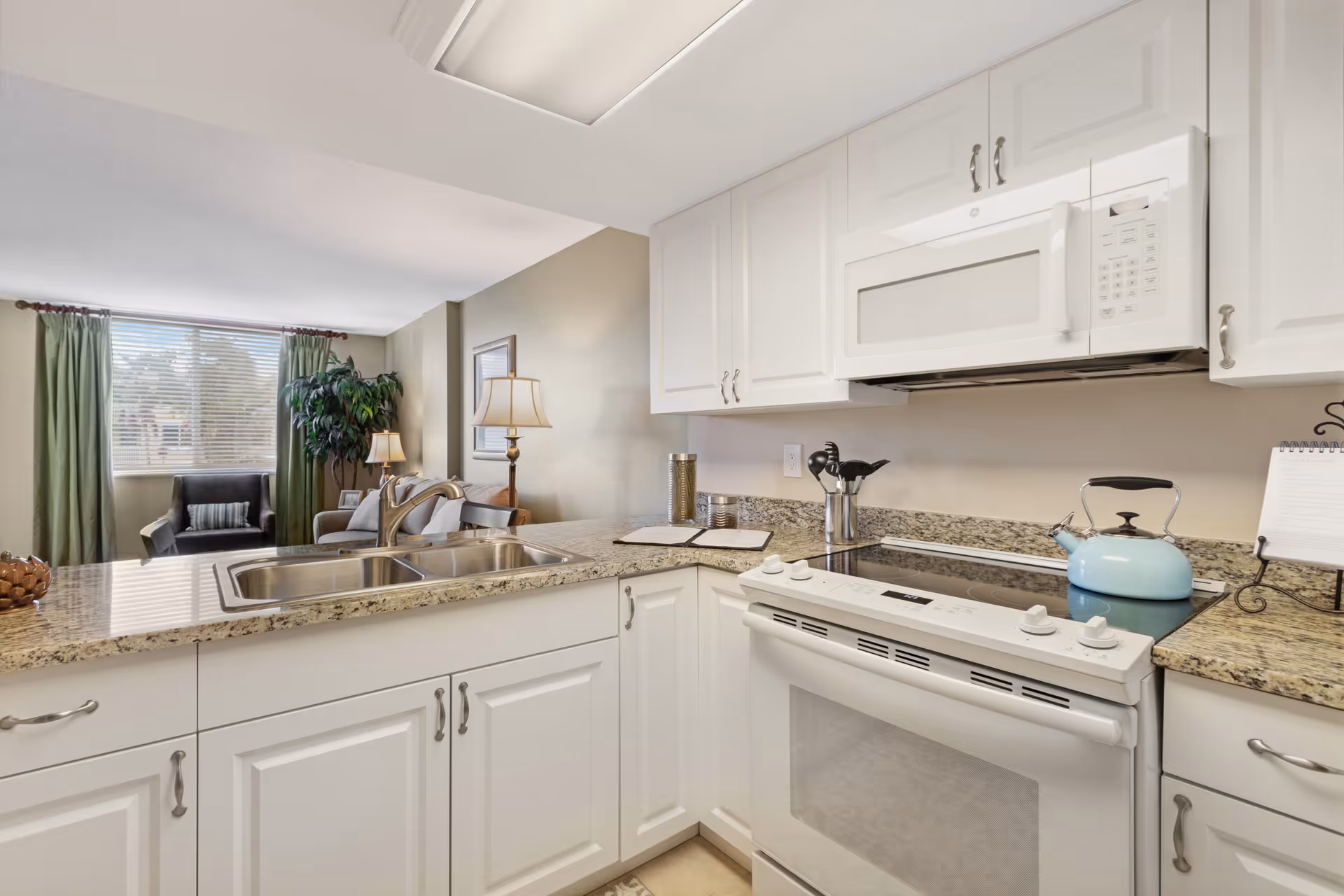 Bright kitchen area with white cabinets, granite countertops, a double sink, a white stove with a blue kettle on it, and a white microwave above. In the background, there is a living room with a window covered by green curtains, a black armchair, a sofa, a floor lamp, and a large potted plant.
