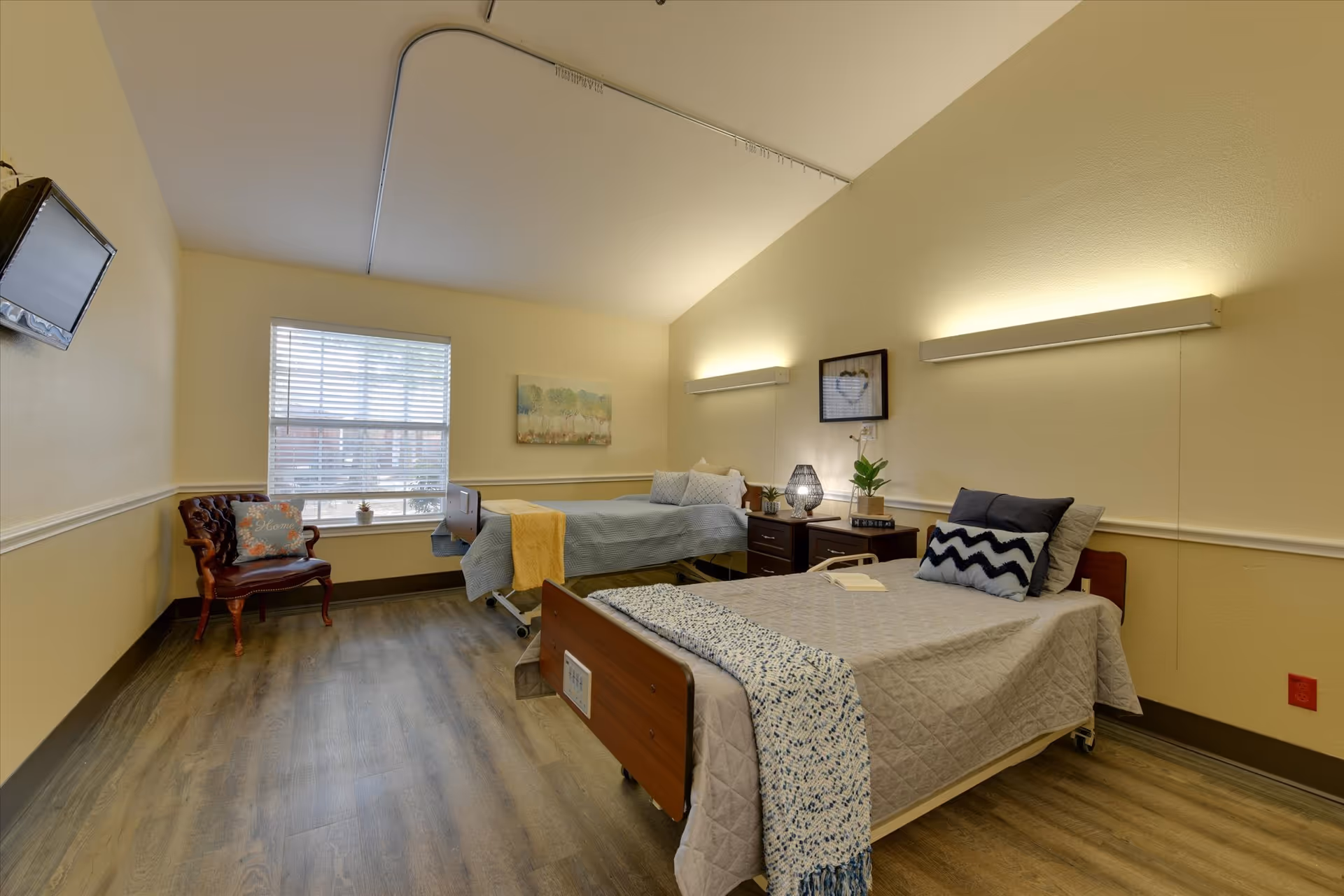Shared senior bedroom with two hospital-style beds, nightstands, and a window letting in natural light.