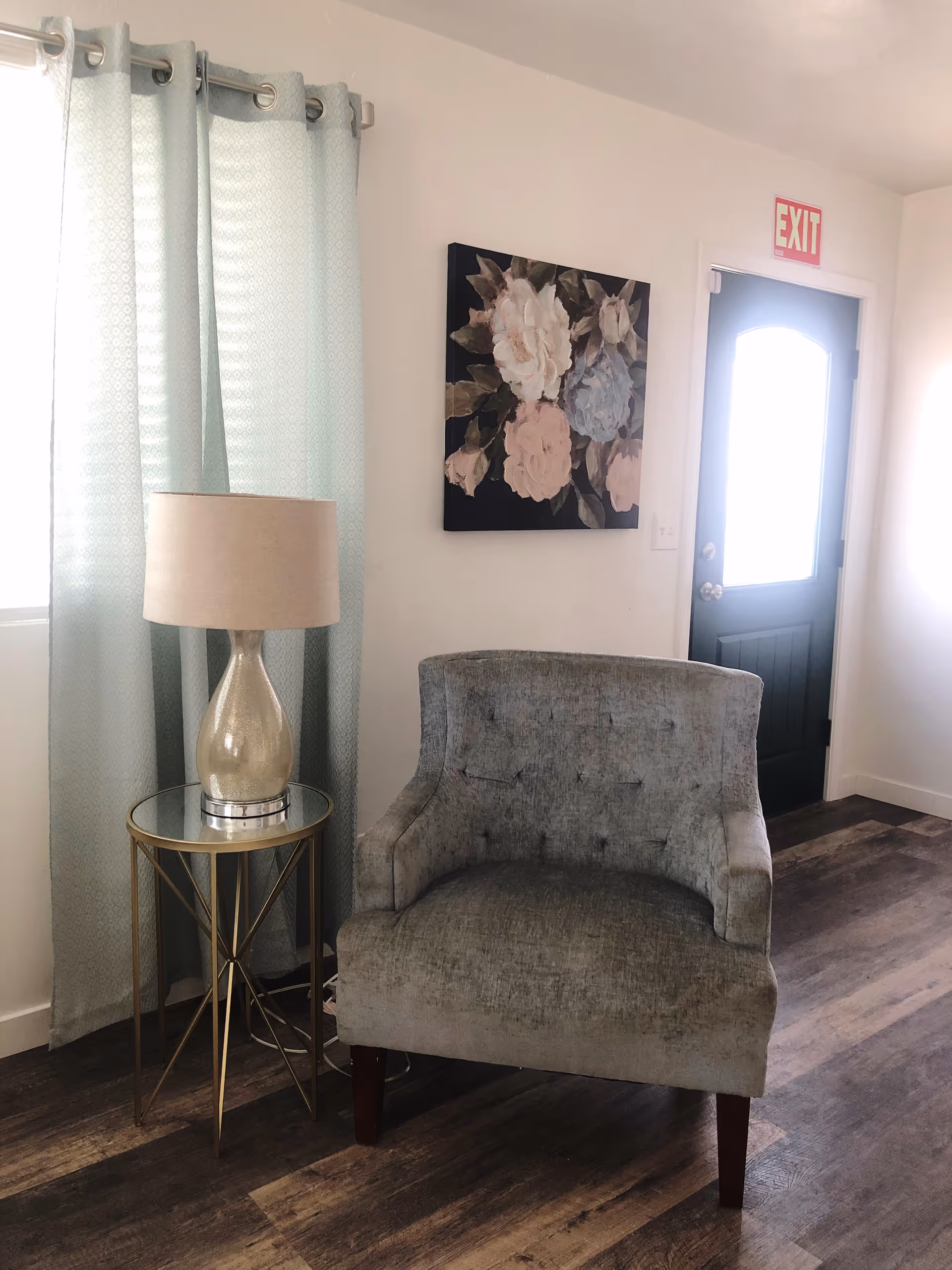 A cozy corner of a room featuring a gray upholstered armchair, a round glass-top side table with a gold metal frame, and a beige table lamp. Light blue curtains hang beside a window, and a floral painting is mounted on the white wall above the chair. A dark-colored door with a window and a red EXIT sign above it is visible in the background. The floor is wood with a natural finish.