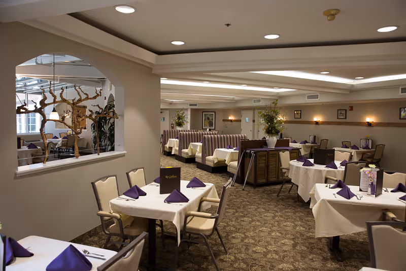Interior view of a senior living facility dining room with tables covered in white tablecloths and purple napkins. The room features beige walls, patterned carpet, booth seating along the back wall, and decorative plants. Soft lighting creates a warm and inviting atmosphere.