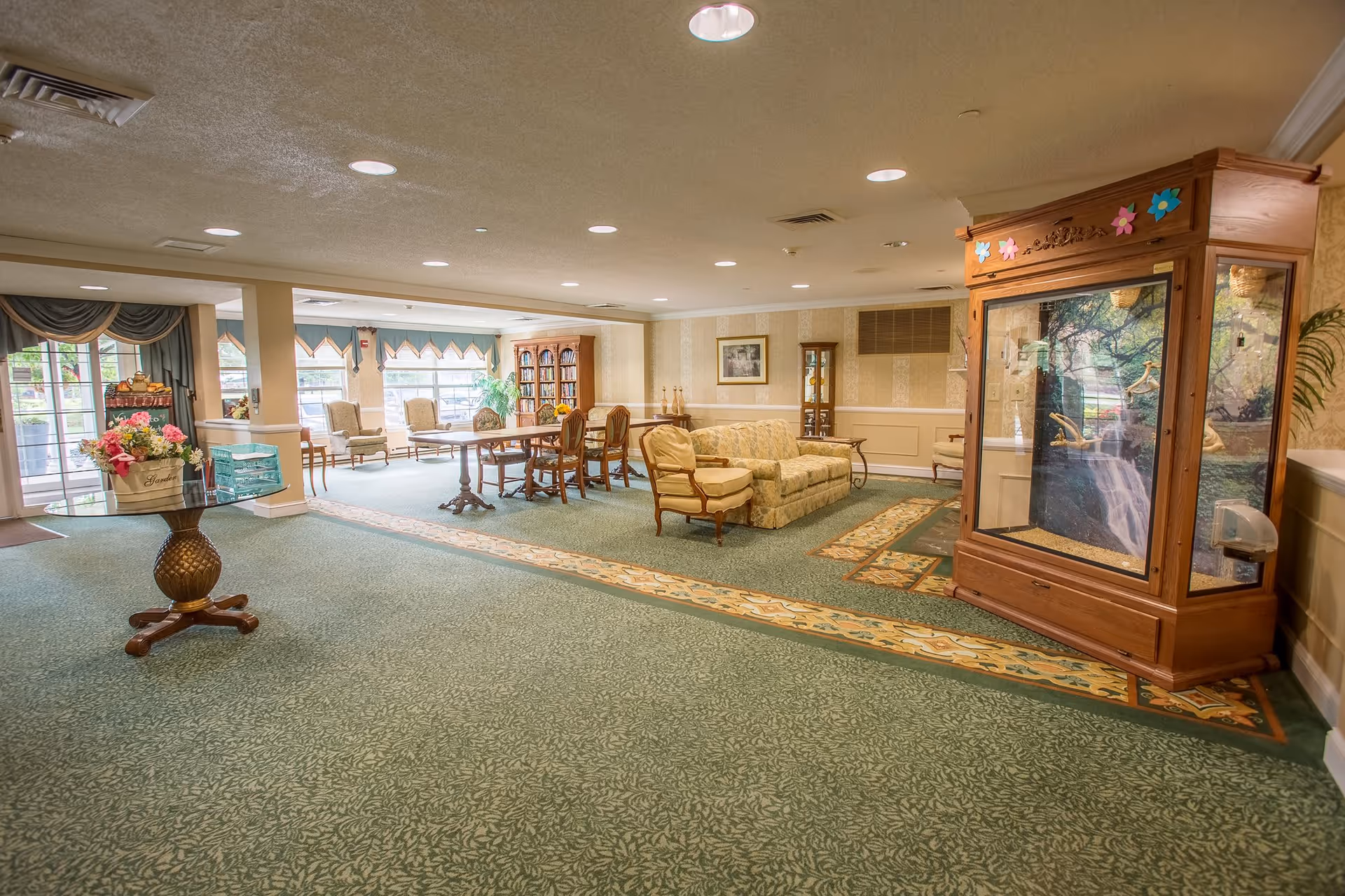 A spacious, well-lit common area in a senior living facility featuring a green patterned carpet, a round table with a flower arrangement, several armchairs, a sofa, a wooden dining table with chairs, a bookshelf, and a large wooden display cabinet with a nature-themed backdrop. Large windows allow natural light to fill the room.