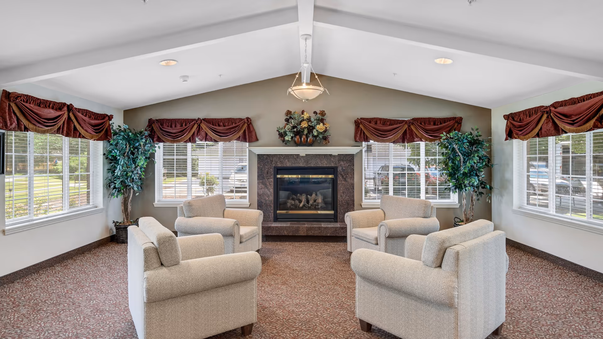 A bright common lounge with four upholstered armchairs arranged around a central fireplace under a vaulted ceiling, flanked by large windows and potted plants.