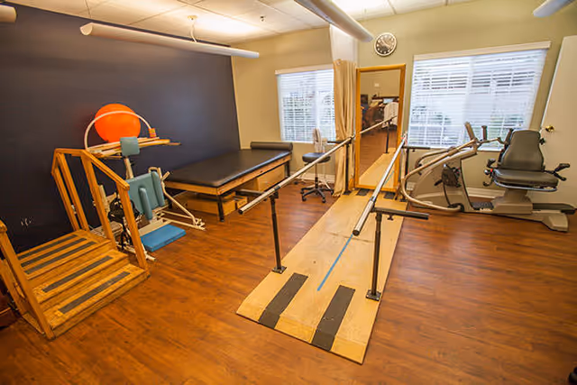 Physical therapy room with wooden stairs, exercise equipment, parallel bars for walking practice, a treatment table, and a recumbent exercise bike. The room has wooden flooring, a large mirror, two windows with blinds, and a clock on the wall.