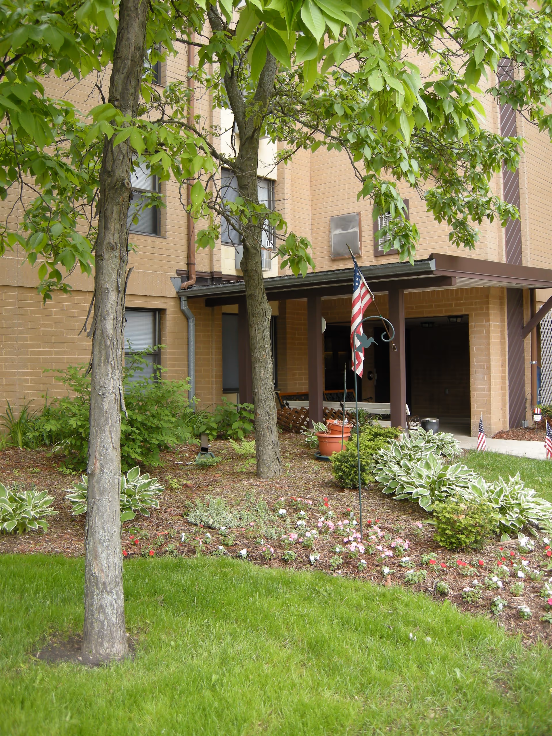 Outdoor garden area with green grass, trees, and flower beds in front of a brick building. An American flag is displayed on a pole in the garden near the building entrance.
