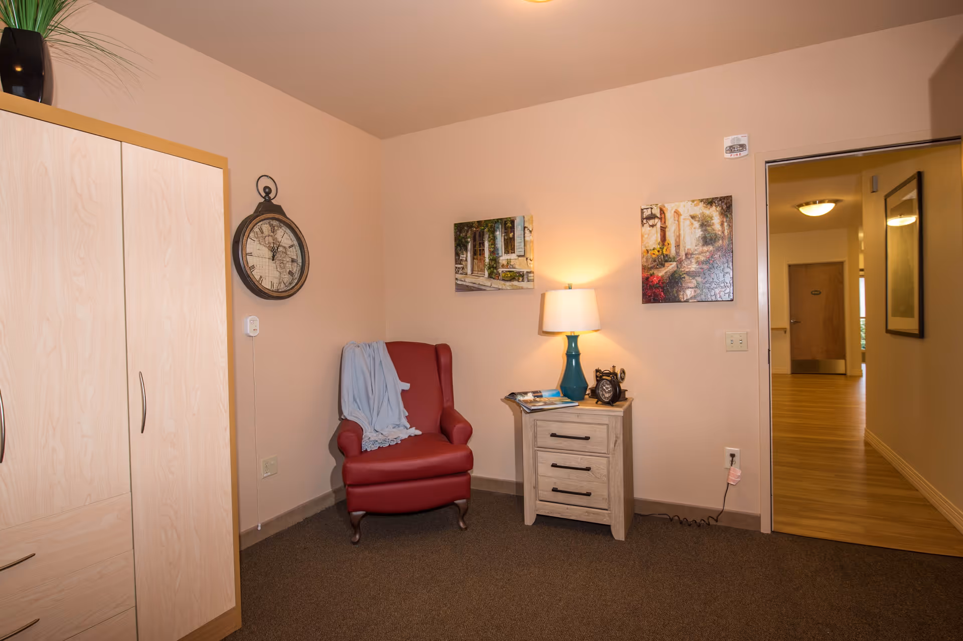 A cozy corner of a room featuring a red armchair with a light blue throw draped over it, a small wooden nightstand with a blue lamp and a vintage clock, two framed paintings on the wall, a large wall clock, and a light wooden wardrobe. The room has beige walls and carpeted flooring, with an open doorway leading to a hallway with wooden flooring.