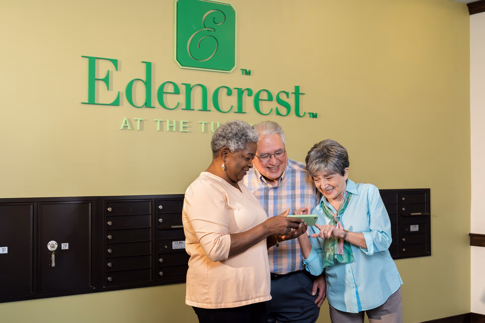 Three elderly people standing together in front of a wall with the Edencrest logo, looking at a smartphone and smiling. Behind them are black mailboxes mounted on the wall.