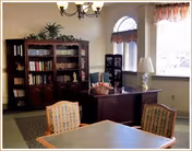 Communal dining/activity room with a table and upholstered chairs in the foreground, bookshelves and a desk near windows and a chandelier overhead.