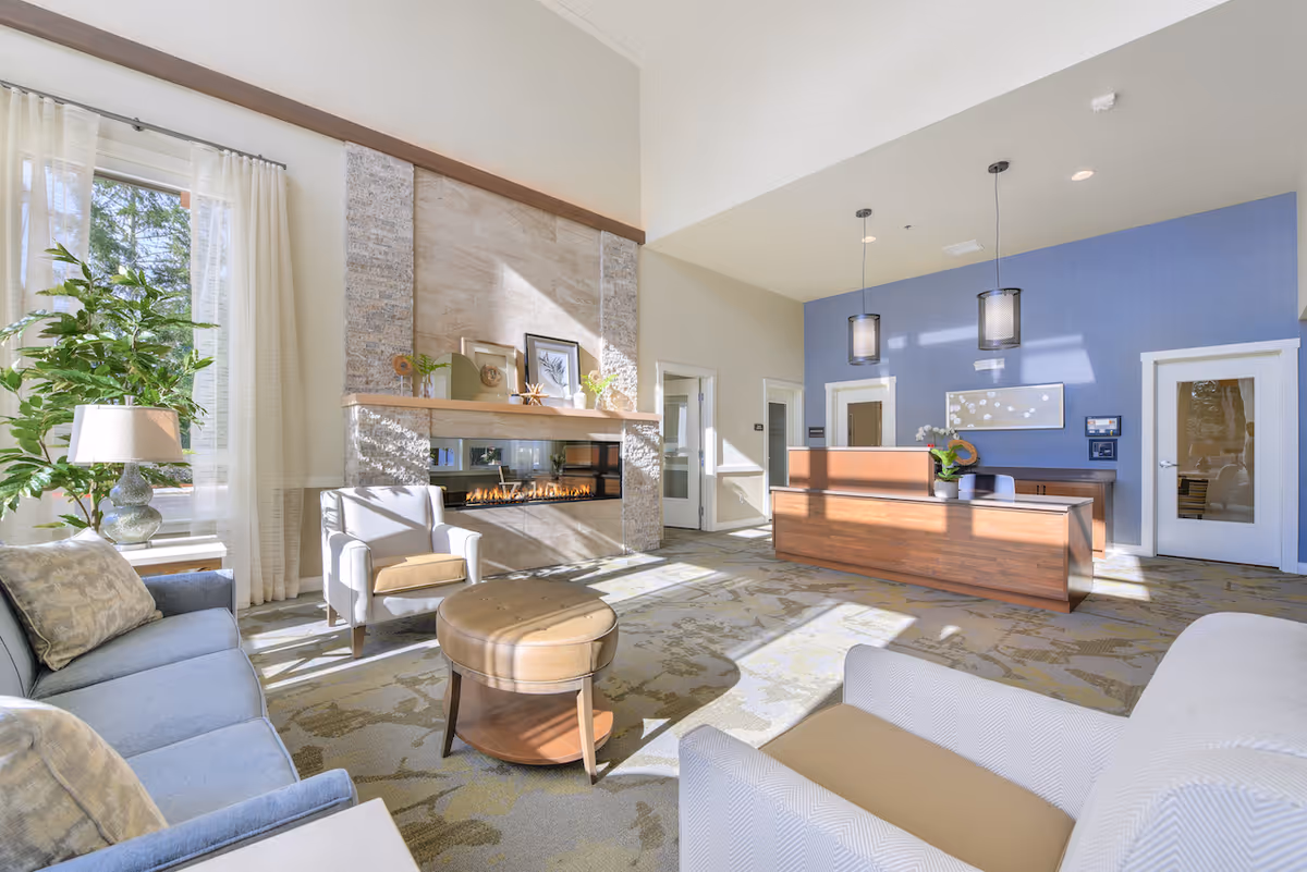 A bright and spacious reception area with a modern wooden front desk, two pendant lights hanging from the ceiling, and a cozy seating area featuring a blue sofa, two armchairs, and a round ottoman. A large window with sheer curtains allows natural light to fill the room. There is a decorative fireplace with framed artwork and plants on the mantel, and a blue accent wall behind the reception desk.