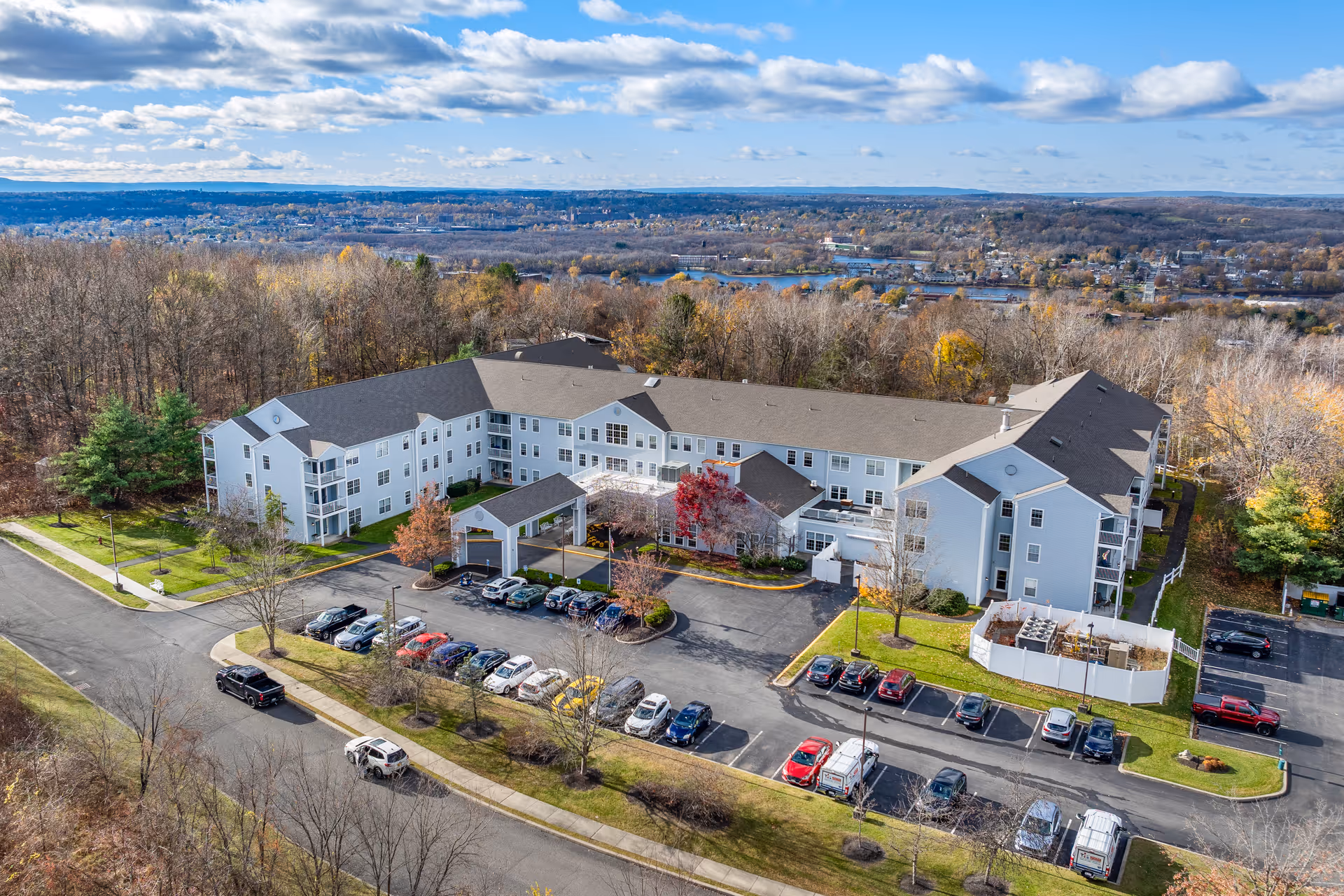 Aerial view of a large, white senior living community building named Diamond Ridge, surrounded by trees with autumn foliage. The building has multiple floors and a covered entrance with a parking lot filled with cars in front. The background shows a distant town and a partly cloudy sky.