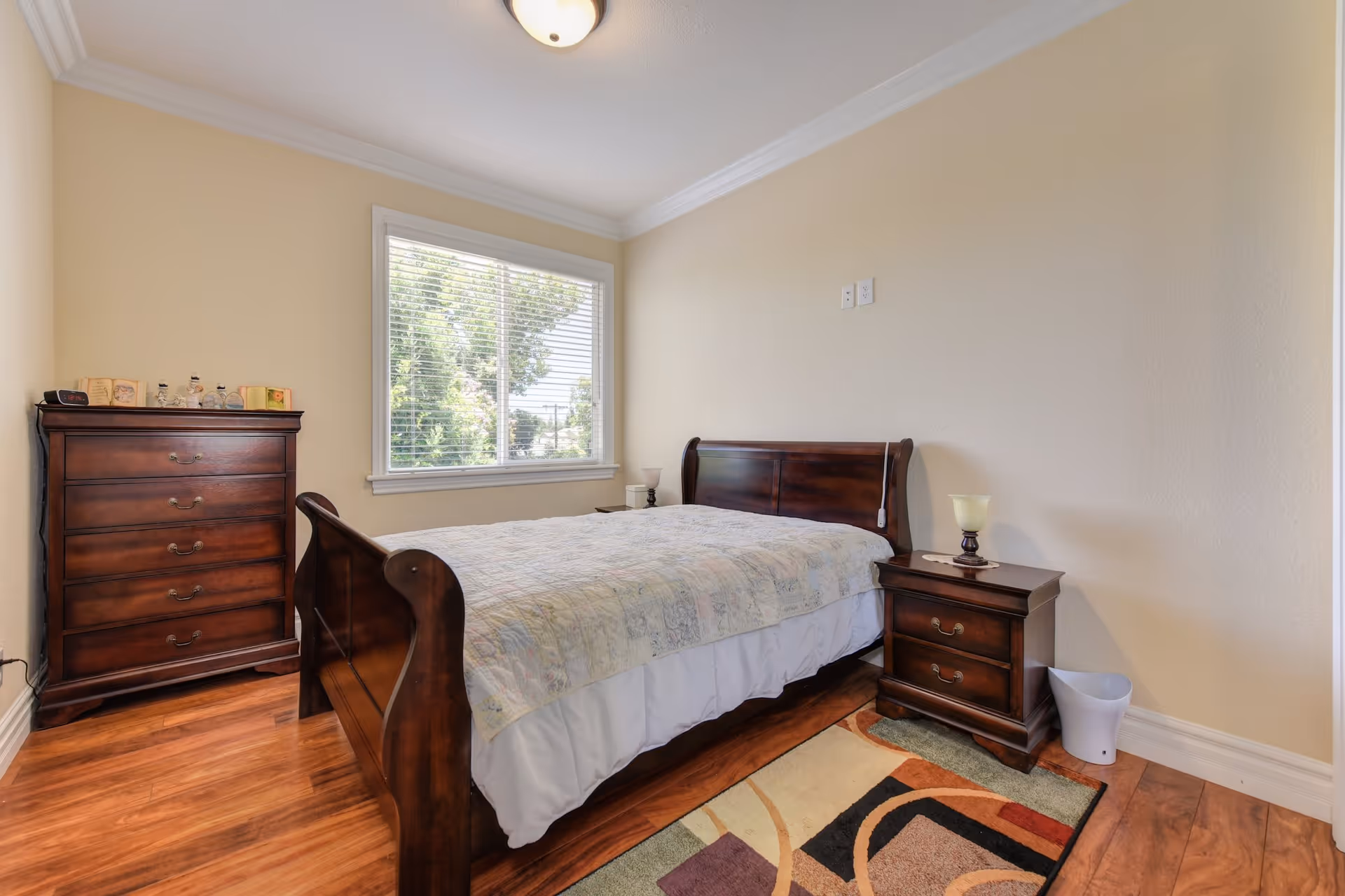 A bedroom with a wooden bed frame and a light-colored quilt on the bed. There is a wooden nightstand with a lamp on the right side of the bed and a wooden chest of drawers against the left wall. A window with white blinds lets in natural light, and a colorful area rug is partially visible on the wooden floor.