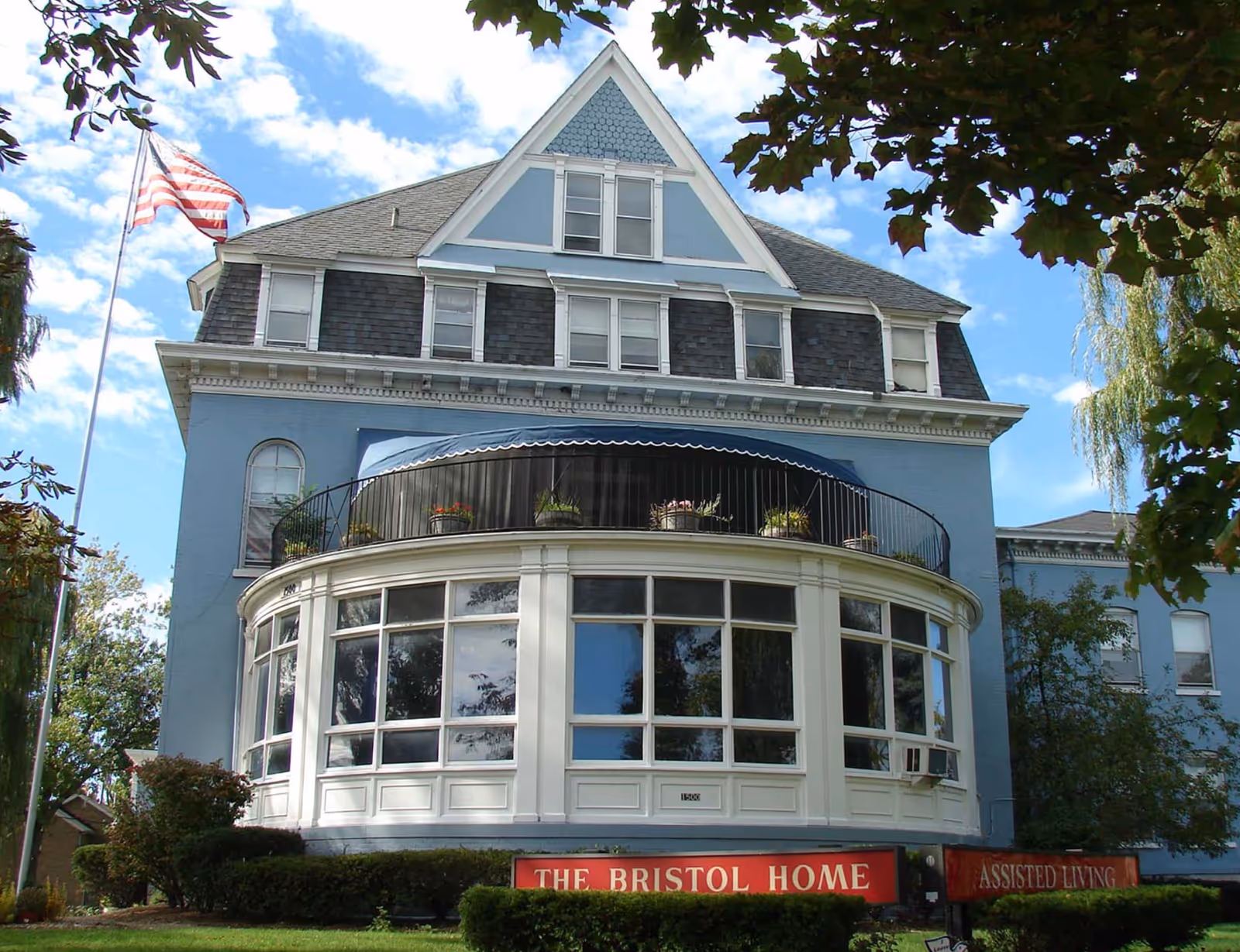 Front exterior of a blue Victorian-style assisted living home with a rounded bay window, balcony, American flag, and a sign reading "The Bristol Home".