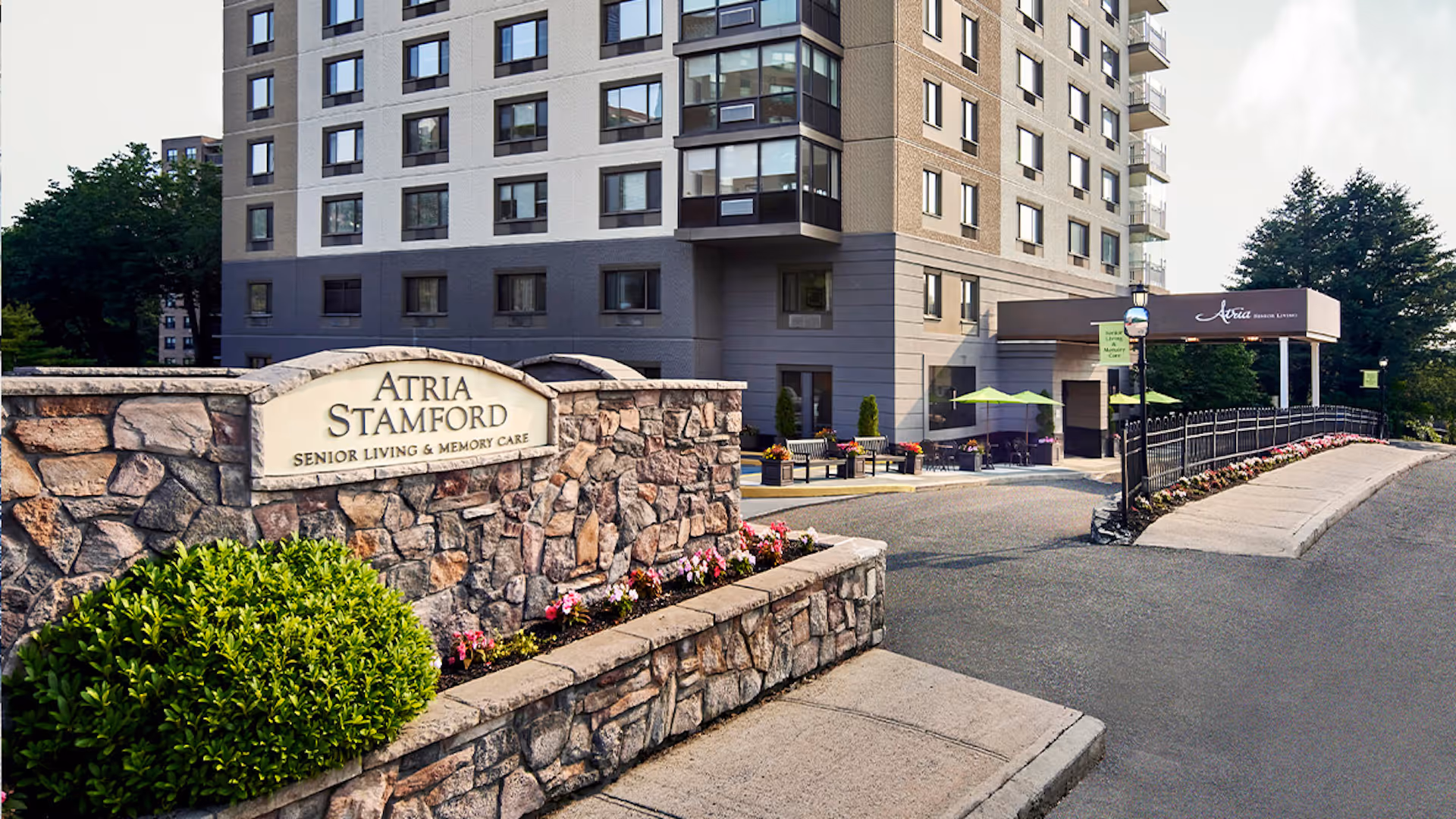 Exterior view of Atria Stamford senior living and memory care facility showing a multi-story building, stone sign with the facility name, landscaped bushes and flowers, and a covered entrance with outdoor seating and green umbrellas.