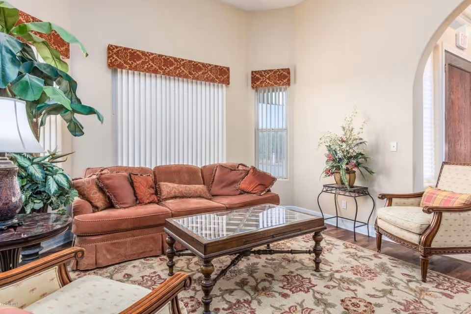 A cozy living room with a rust-colored sofa adorned with multiple cushions, two upholstered armchairs with wooden frames, a glass-top wooden coffee table, a patterned area rug, a side table with a lamp and large green plants, and a small table with a floral arrangement near an arched doorway.