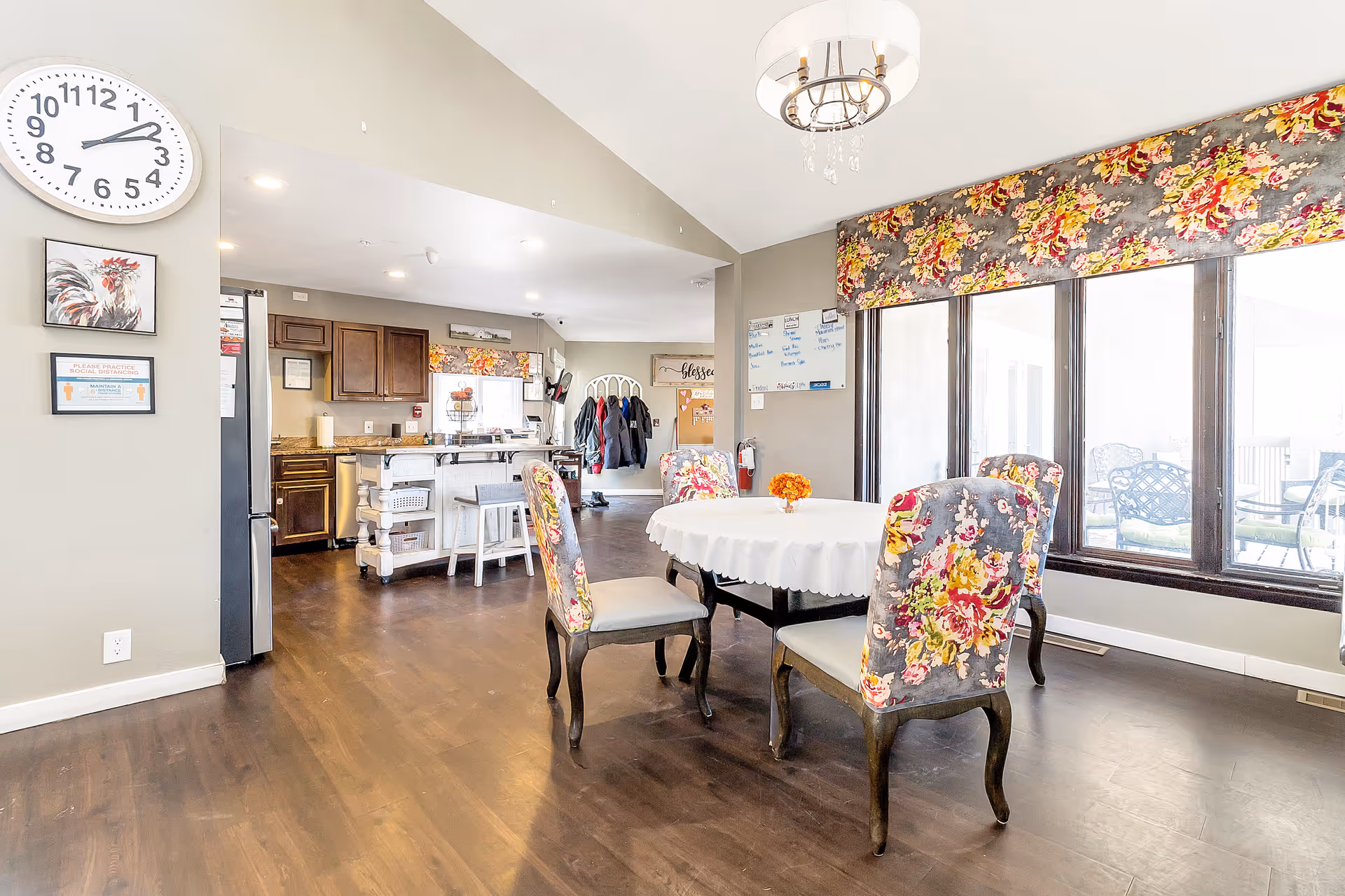 Bright dining area with a round table covered with a white tablecloth and four floral upholstered chairs. Large windows with floral valances let in natural light. In the background, a kitchen area with wooden cabinets, a refrigerator, and a kitchen island with stools is visible. A clock and framed pictures hang on the wall, and a chandelier hangs from the ceiling.