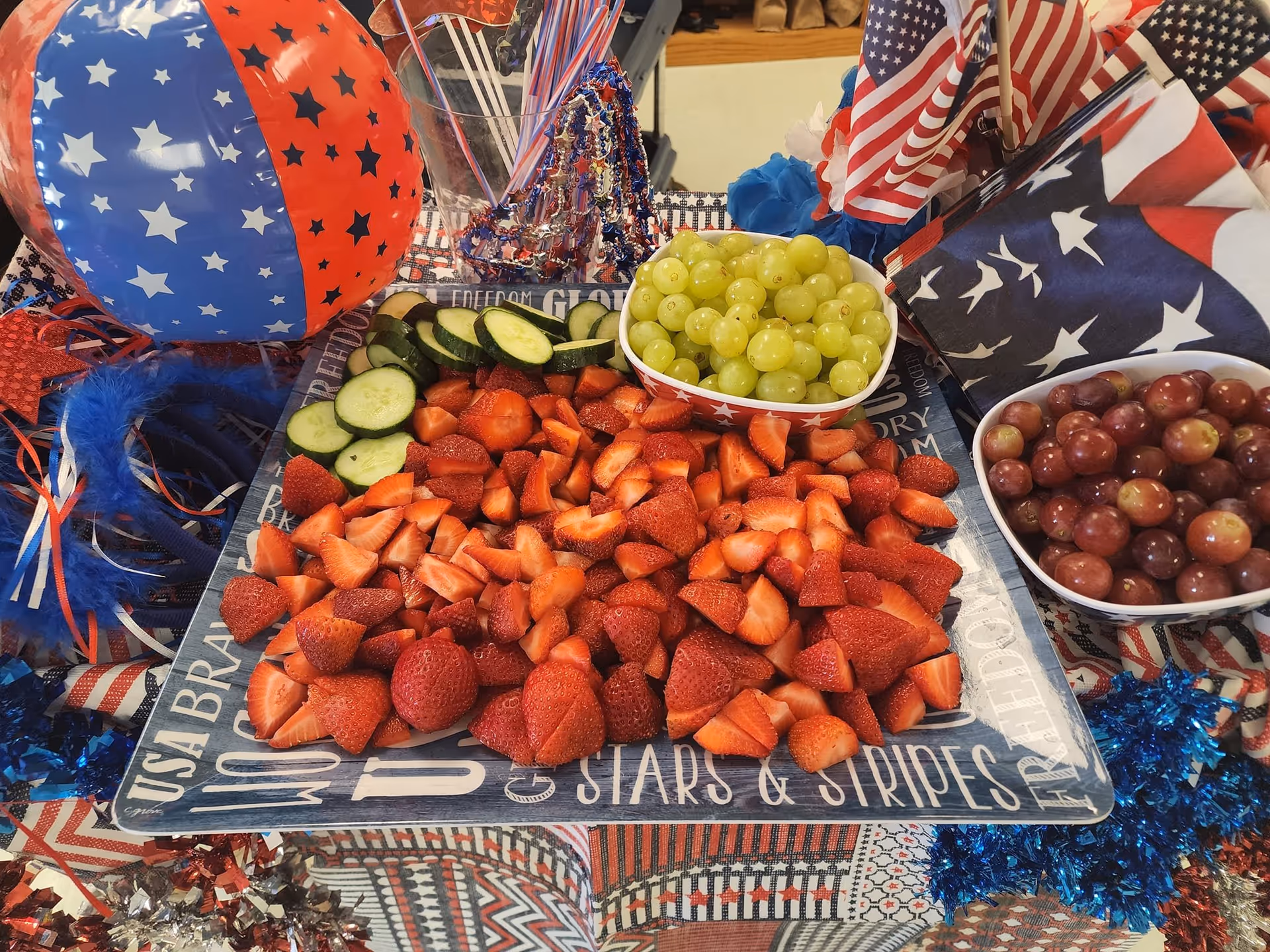 A patriotic-themed table setting featuring a platter of sliced strawberries, cucumber slices, a bowl of green grapes, and a bowl of red grapes. The table is decorated with red, white, and blue items including a star-patterned beach ball, small American flags, and festive garlands.