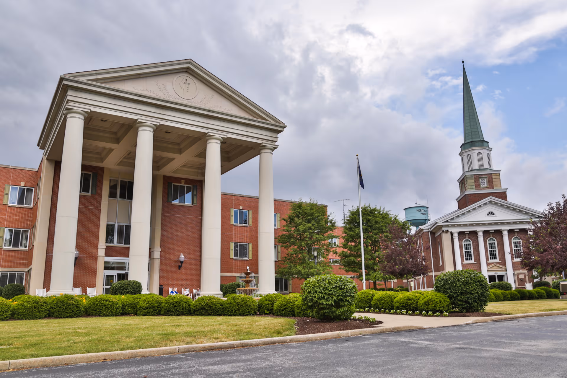 Exterior front view of Heritage Pointe of Warren showing a brick building with a tall columned portico, landscaped grounds, and a steeple.