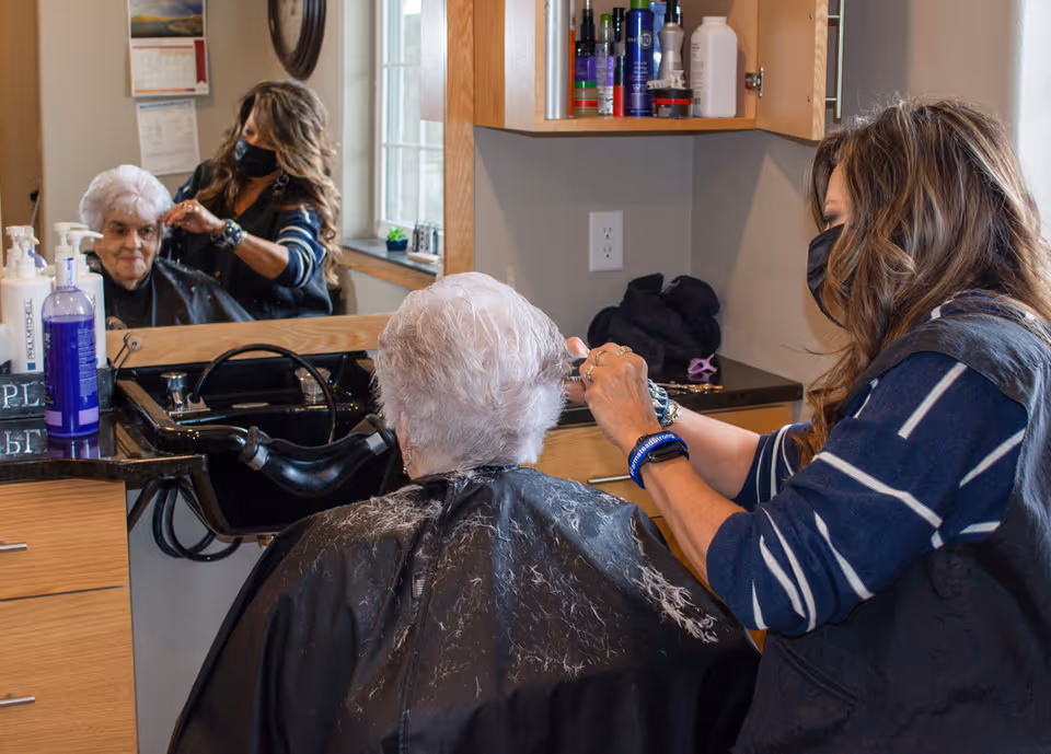 A hairstylist wearing a mask is cutting the hair of an elderly woman seated in a salon chair with a black cape. The scene is reflected in a large mirror in front of them, showing the elderly woman's face and the hairstylist working. Various hair care products are visible on a shelf and counter nearby.