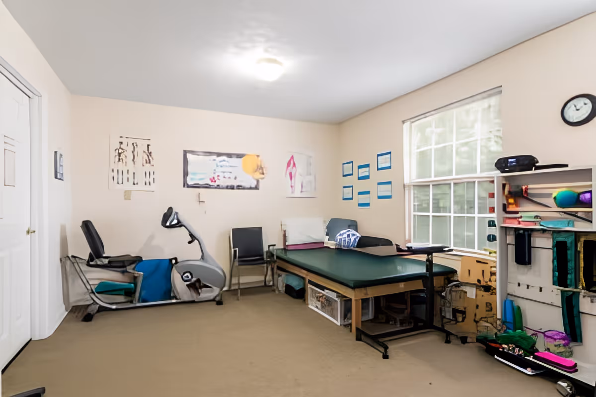 A therapy or exercise room with a recumbent exercise bike, a padded treatment table, chairs, and shelves with various supplies. The room has beige walls, a large window with blinds, and a clock on the wall.