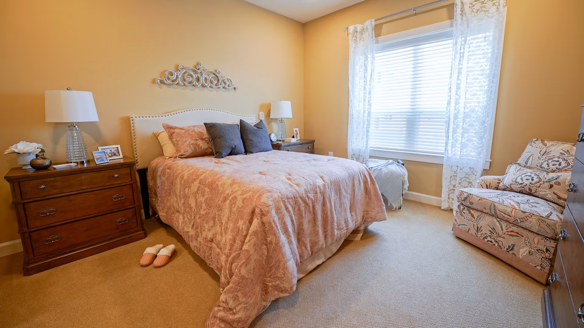 A cozy bedroom with a large bed covered in a pink patterned comforter and multiple pillows. There are two wooden nightstands on either side of the bed, each with a lamp and decorative items. A pair of slippers is placed on the carpeted floor next to the bed. A window with white blinds and sheer floral curtains lets in natural light. A floral upholstered armchair is positioned near the window.