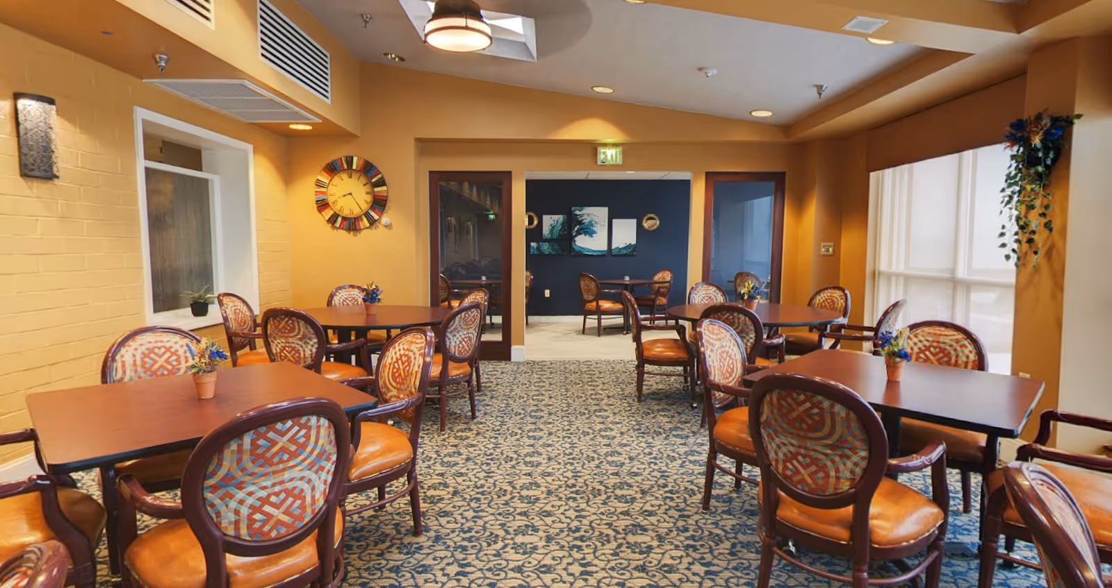 Interior view of a dining area in Carroll Lutheran Village with multiple wooden tables and cushioned chairs arranged neatly on a patterned carpet. The walls are painted warm yellow and decorated with a colorful clock and some artwork. Large windows on the right side allow natural light to brighten the room, and small potted plants are placed on the tables.