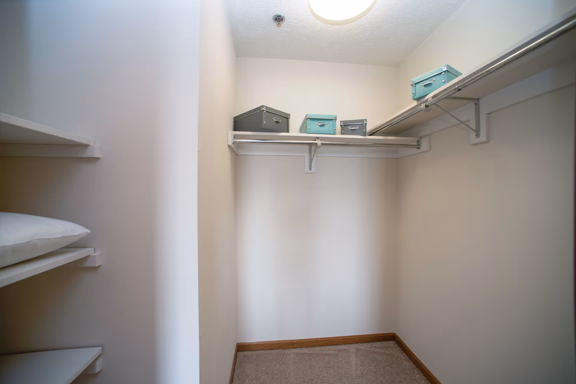 A small walk-in closet with beige walls and carpeted floor. There are white shelves on the left side with a pillow on one shelf, and a white hanging rod with four storage boxes in teal and gray colors on the right side near the ceiling. A round ceiling light illuminates the space.